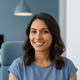 Headshot of a smiling South Asian woman in a slate blue top, with a soft dusty blue and white background.